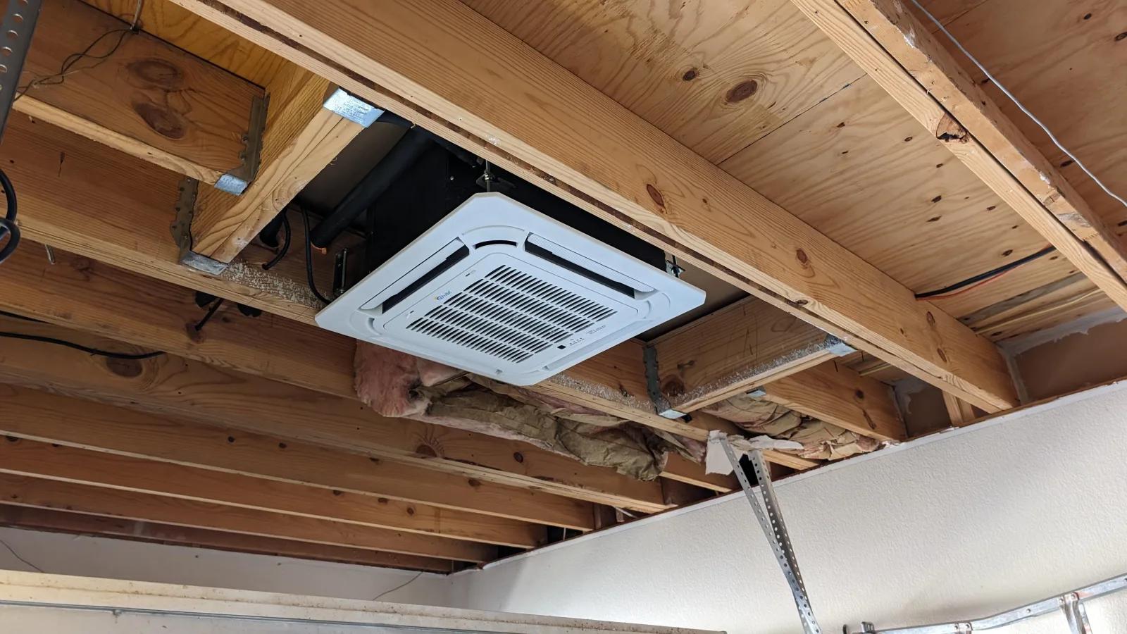 In ceiling mini split — recessed ceiling cassette installed flush between exposed floor joists, with its four-way airflow grille facing down into the room